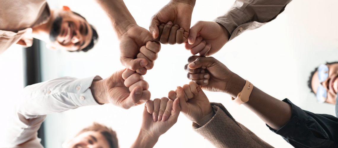 Teamwork, Power And Partnership Concept. Below view of multucultural group of smiling people making fist bump standing in circle. Workers doing fist pump together celebrating good deal