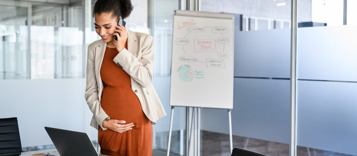 Smiling pregnant businesswoman touching belly while using laptop and talking over phone in modern office. Cheerful pregnant entrepreneur talking on phone while working with copy space. Beautiful african american woman working on laptop and talking on mobile phone.